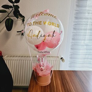 May include: A clear balloon shaped like a hot air balloon with pink and white balloons inside. The balloon has gold lettering that says "Welcome to the World Adiyah" and a pink ribbon with a tag that says "Poshy Giftbox". The balloon is sitting on a table with a pink gift box with a pink teddy bear inside.