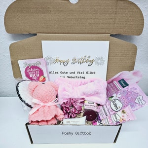 Peut inclure: Une bo&icirc;te cadeau rose remplie de divers articles, dont un ours en peluche rose, un livre intitul&eacute; "Jegurite Schon Dass Es Dich Gibt", un bandeau rose, une bougie et des produits de bain. La bo&icirc;te est &eacute;tiquet&eacute;e "Poshy Giftbox" et comporte une carte avec le texte "Happy Birthday Alles Gute und Viel Gl&uuml;ck zum Geburtstag."