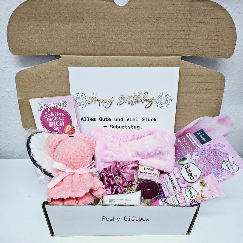 Peut inclure: Une bo&icirc;te cadeau rose remplie de divers articles, dont un ours en peluche rose, un livre intitul&eacute; "Jegurite Schon Dass Es Dich Gibt", un bandeau rose, une bougie et des produits de bain. La bo&icirc;te est &eacute;tiquet&eacute;e "Poshy Giftbox" et comporte une carte avec le texte "Happy Birthday Alles Gute und Viel Gl&uuml;ck zum Geburtstag."