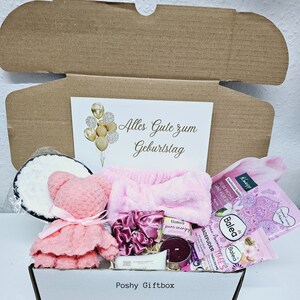 Peut inclure: Une bo&icirc;te cadeau remplie de divers articles, dont un ours en peluche rose, un bandeau rose, une bombe de bain blanche, une poudre de bain rose, une bougie, une bouteille de lotion pour le corps et une carte qui dit "Alles Gute zum Geburtstag" (Joyeux anniversaire).