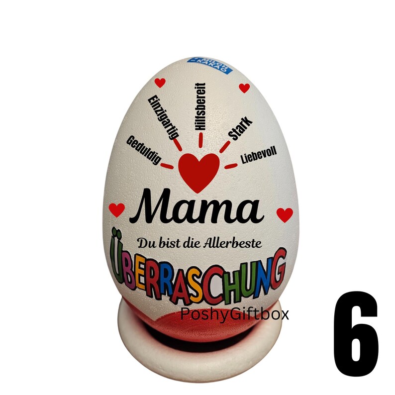 May include: A white egg-shaped decorative item with the word "Mama" in black script, a red heart, and the phrase "Du bist die Allerbest" in black. The egg has the word "&Uuml;berraschung" in colorful letters. It sits on a white and red base.