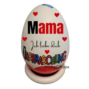 May include: White egg-shaped decorative item with the word "Mama" in red, "Ich liebe dich" in black script, and "&Uuml;BERRASCHUNG" in colorful letters. Red heart accents are scattered across the surface. The egg rests on a white and red base.