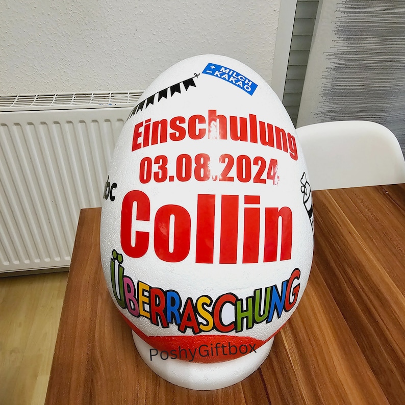Peut inclure: Conteneur blanc en forme d'&oelig;uf avec le texte "Einschulung 03.08.2024 Collin &Uuml;berraschung" et le logo "PoshyGiftbox".