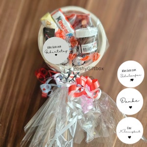 May include: A white bowl filled with various candies and chocolate bars, wrapped in clear cellophane and tied with red and white ribbon. The bowl is attached to a wooden stick and decorated with white circular tags with black text, including "Alles Gute zum Geburtstag", "Alles Gute zum Schulanfan", "Danke", and "Eine Kleinigkeit".