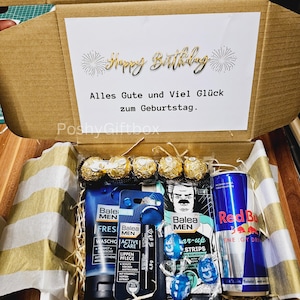 May include: A brown cardboard box with a white card inside that says "Happy Birthday" in gold lettering. The box is open and contains a variety of items, including a can of Red Bull, a tube of face wash, a tube of lip balm, a pack of face masks, a pack of razor blades, and a few chocolate candies.