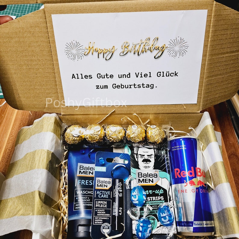May include: A brown cardboard box with a white card inside that says "Happy Birthday" in gold lettering. The box is open and contains a variety of items, including a can of Red Bull, a tube of face wash, a tube of lip balm, a pack of face masks, a pack of razor blades, and a few chocolate candies.