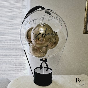 May include: A clear balloon hot air balloon with gold balloons inside. The balloon is decorated with black ribbon and a black box at the bottom. The balloon has the text "Happy 30th Birthday Stephanie" written on it.