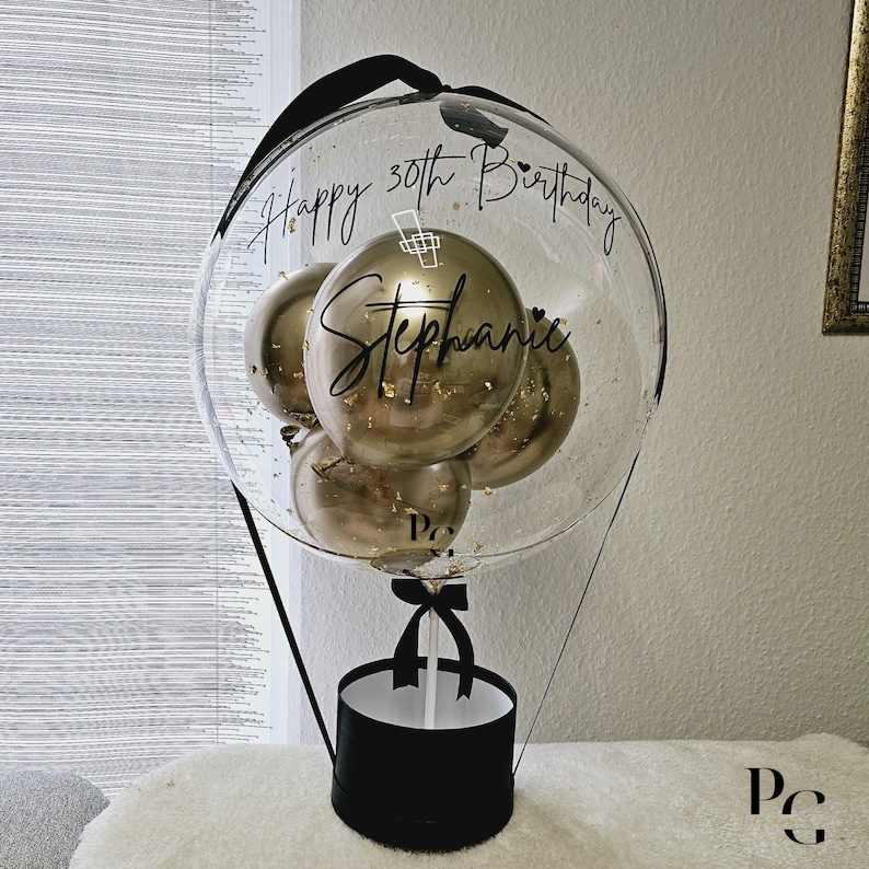 May include: A clear balloon hot air balloon with gold balloons inside. The balloon is decorated with black ribbon and a black box at the bottom. The balloon has the text "Happy 30th Birthday Stephanie" written on it.