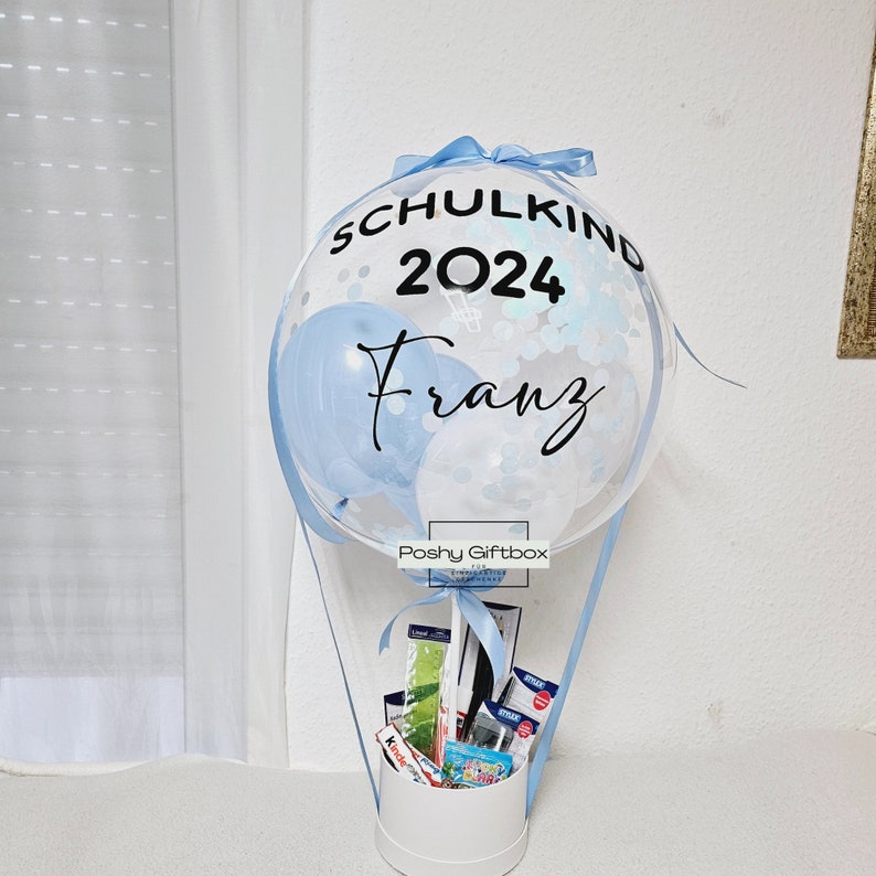 May include: A blue and white balloon hot air balloon with the text "Schulkind 2024 Franz" on it. The balloon is filled with confetti and is attached to a white box with a blue ribbon. The box contains school supplies.