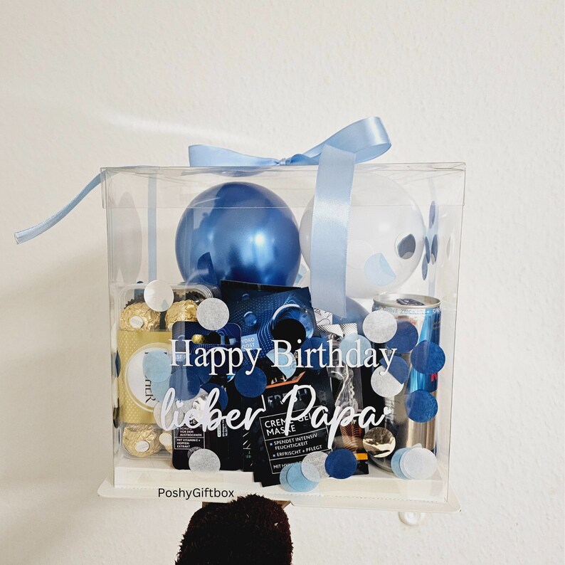 May include: A clear plastic box filled with blue and white balloons, chocolate candies, a can of energy drink, and confetti. The box is tied with a blue ribbon and has the text "Happy Birthday lieber Papa" written on it.