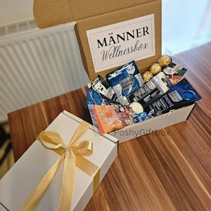 May include: A white gift box with a gold ribbon and a brown box filled with various items, including toiletries, a candle, and chocolates. The brown box has a white label that reads "M&Auml;NNER Wellnessbox".
