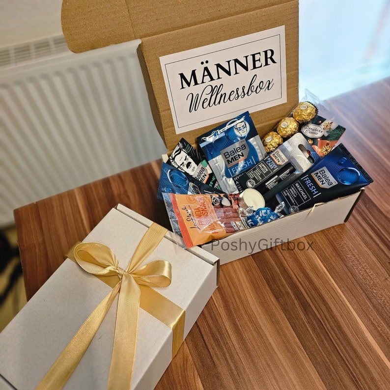 May include: A white gift box with a gold ribbon and a brown box filled with various items, including toiletries, a candle, and chocolates. The brown box has a white label that reads "M&Auml;NNER Wellnessbox".