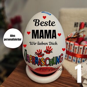 Pode incluir: Uma caixa de presente branca em forma de ovo com as palavras "Beste MAMA" e "Wir lieben Dich" em preto. O ovo é decorado com corações vermelhos e a palavra "Überraschung" em letras coloridas. A base é branca.