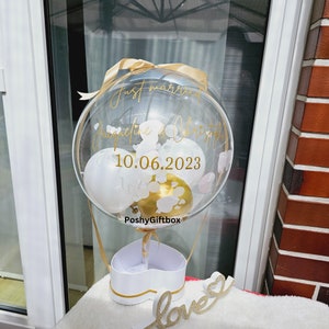 May include: A clear balloon with a gold ribbon and white balloons inside. The balloon has the text "Just married Jacqueline & Christopher 10.06.2023" and "PoshyGiftbox" printed on it. The balloon is sitting on top of a white heart-shaped box with a gold trim. The box is sitting on a white surface with a gold "love" sign.