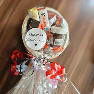 May include: A white gift basket filled with chocolate bars, candy, and a small jar of Nutella. The basket is wrapped in clear cellophane and tied with red, white, and silver ribbon. A white circle sticker with the text "Alles Gute zum Schulanfang" is attached to the basket.