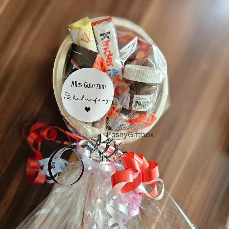 May include: A white gift basket filled with chocolate bars, candy, and a small jar of Nutella. The basket is wrapped in clear cellophane and tied with red, white, and silver ribbon. A white circle sticker with the text "Alles Gute zum Schulanfang" is attached to the basket.