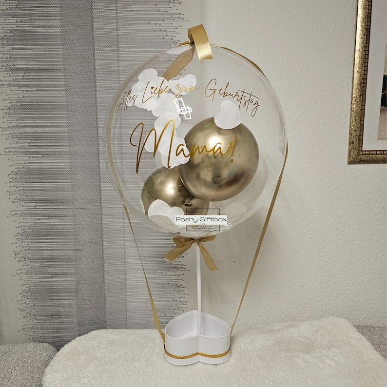 May include: A clear balloon shaped like a hot air balloon with gold ribbon and a white heart-shaped base. Inside the balloon are three gold balloons and white paper confetti. The balloon has the text "Alles Liebe zum Geburtstag Mama!" written on it.