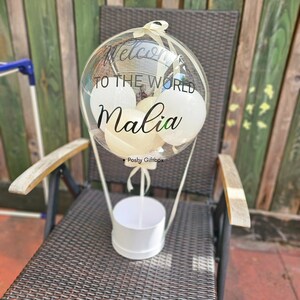 May include: A clear balloon with white balloons inside, a white ribbon, and the text "Welcome to the world Malia" in black lettering. The balloon is attached to a white box with the text "Poshy Giftbox" in black lettering.
