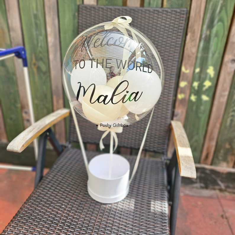 May include: A clear balloon with white balloons inside, a white ribbon, and the text "Welcome to the world Malia" in black lettering. The balloon is attached to a white box with the text "Poshy Giftbox" in black lettering.