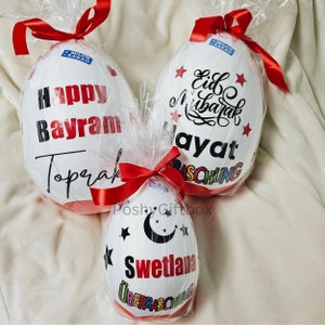May include: Three white chocolate eggs wrapped in clear plastic with red ribbons. The eggs are decorated with black text and colorful stars. The text on the eggs reads "Happy Bayram Toprak", "Eid Mubarak Ayat", and "Svetlana".
