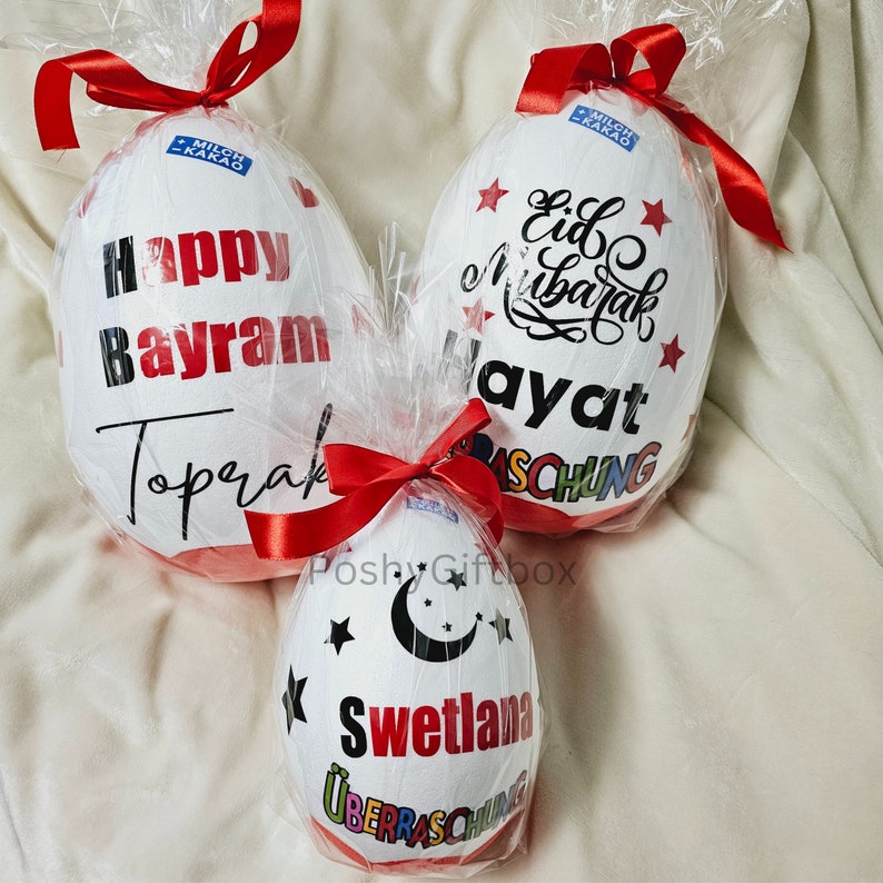 May include: Three white chocolate eggs wrapped in clear plastic with red ribbons. The eggs are decorated with black text and colorful stars. The text on the eggs reads "Happy Bayram Toprak", "Eid Mubarak Ayat", and "Svetlana".