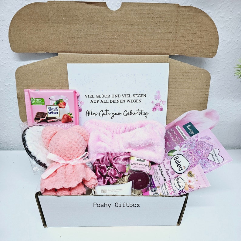 Peut inclure: Une bo&icirc;te cadeau rose remplie de divers articles, dont un ours en peluche rose, un bandeau rose, une barre de chocolat, une bougie et un produit de bain. La bo&icirc;te est &eacute;tiquet&eacute;e "Poshy Giftbox" et comprend une carte avec le texte "Viel Gl&uuml;ck und viel Segen auf all deinen Wegen. Alles Gute zum Geburtstag".