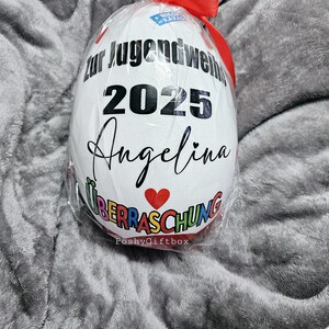 May include: A white, egg-shaped gift wrapped in clear cellophane and tied with a red ribbon. The egg features black text that reads "zur Jugendweihe 2025 Angelina" with a red heart and the word "&Uuml;berraschung".