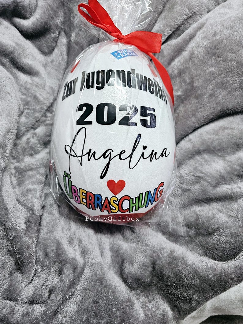 May include: A white, egg-shaped gift wrapped in clear cellophane and tied with a red ribbon. The egg features black text that reads "zur Jugendweihe 2025 Angelina" with a red heart and the word "&Uuml;berraschung".