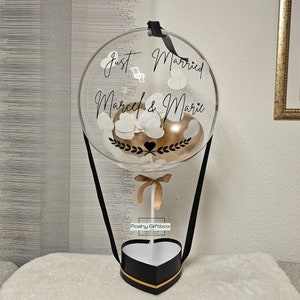May include: A clear balloon hot air balloon with white confetti and a gold balloon inside. The balloon is tied to a black and gold heart-shaped box with the text "Poshy Giftbox" on the side. The balloon has the text "Just Married Marcel & Marie" written on it.
