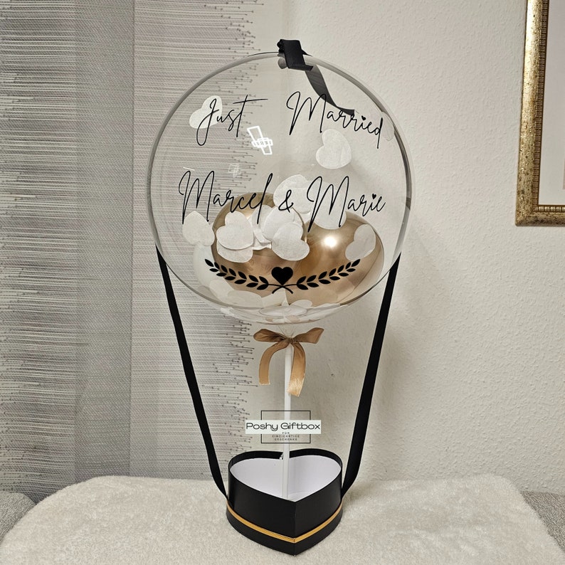 May include: A clear balloon hot air balloon with white confetti and a gold balloon inside. The balloon is tied to a black and gold heart-shaped box with the text "Poshy Giftbox" on the side. The balloon has the text "Just Married Marcel & Marie" written on it.