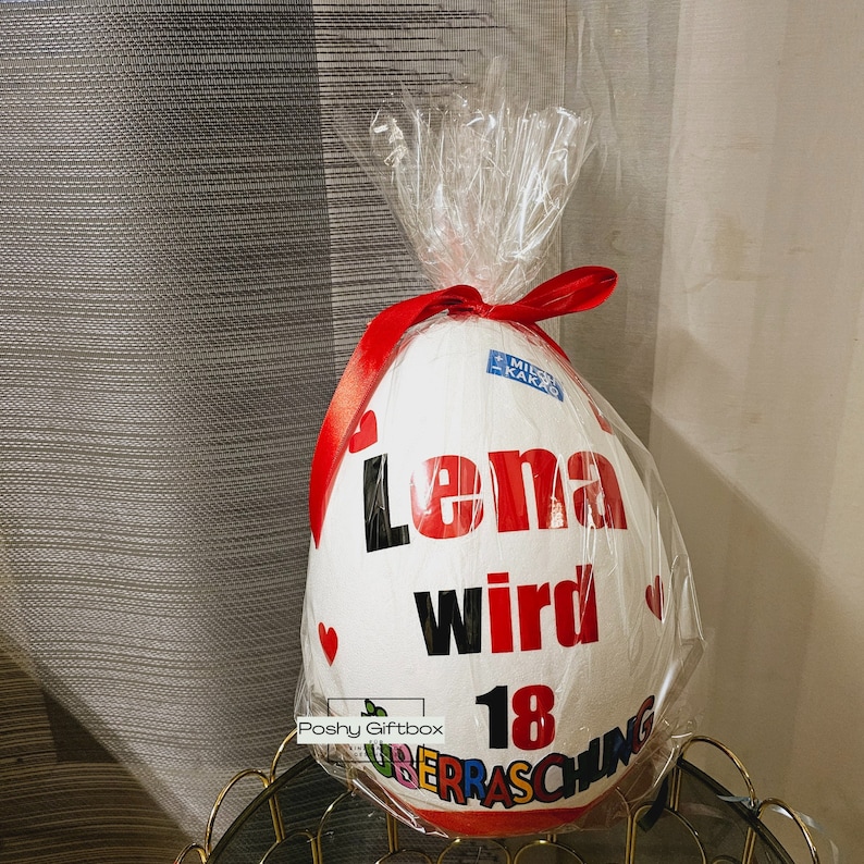 Może przedstawiać: Duży, biały prezent w kształcie jaja, owinięty przezroczystym celofanem i zawiązany czerwoną wstążką. Na jajku widnieją słowa "Lena wird 18 &Uuml;berraschung" wydrukowane w kolorze czerwonym i czarnym. Widoczna jest mała niebiesko-biała etykieta.