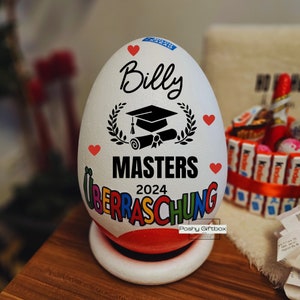 Może przedstawiać: Biały pojemnik w kształcie jajka z nadrukiem czapki absolwenta, wieńca laurowego i zwoju. Na pojemniku widnieje napis "Billy Masters 2024". Pojemnik jest ozdobiony czerwonymi sercami i napisem "&Uuml;berraschung" w kolorach tęczy.