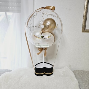 May include: A clear balloon with a heart-shaped box at the bottom. The balloon has a gold ribbon and three balloons inside: one gold, one white, and one white with gold leaves. The balloon has the text "Just Married Anna & Richard" written on it.