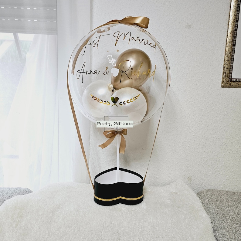 May include: A clear balloon with a heart-shaped box at the bottom. The balloon has a gold ribbon and three balloons inside: one gold, one white, and one white with gold leaves. The balloon has the text "Just Married Anna & Richard" written on it.