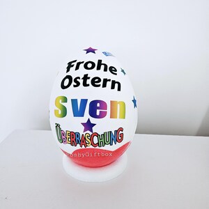 Op de afbeelding: Een wit paasei met een regenboogontwerp en de tekst "Frohe Ostern Sven &Uuml;berraschung" erop. Het ei staat op een rode en witte standaard.