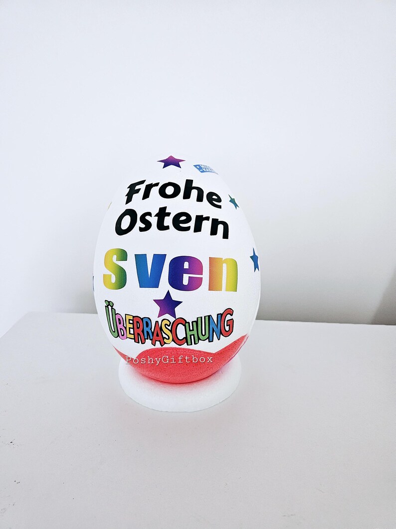 Op de afbeelding: Een wit paasei met een regenboogontwerp en de tekst "Frohe Ostern Sven &Uuml;berraschung" erop. Het ei staat op een rode en witte standaard.