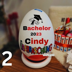 Może przedstawiać: Biały pojemnik w kształcie jajka z wzorem czapki absolwenta i tekstem "Bachelor 2023 Cindy" w czarnych i czerwonych literach. Pojemnik ma kolorowy wz&oacute;r z tekstem "Ueberraschung" w kolorach tęczy. Pojemnik stoi na białej podstawie.