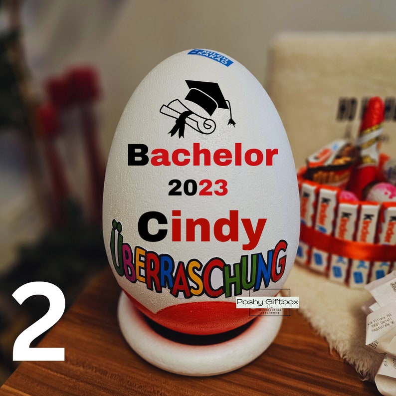 Może przedstawiać: Biały pojemnik w kształcie jajka z wzorem czapki absolwenta i tekstem "Bachelor 2023 Cindy" w czarnych i czerwonych literach. Pojemnik ma kolorowy wz&oacute;r z tekstem "Ueberraschung" w kolorach tęczy. Pojemnik stoi na białej podstawie.