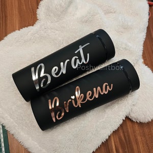 May include: Two black insulated tumblers with personalized names. The tumbler on the left has the name "Berat" in silver lettering. The tumbler on the right has the name "Brikena" in rose gold lettering.