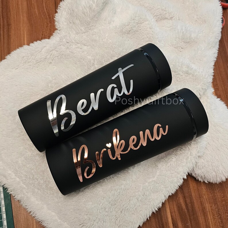 May include: Two black insulated tumblers with personalized names. The tumbler on the left has the name "Berat" in silver lettering. The tumbler on the right has the name "Brikena" in rose gold lettering.