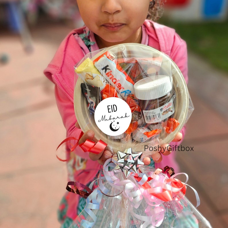 May include: A clear plastic bag filled with assorted candies and chocolate bars, including Kinder chocolate, Nutella, and other treats. The bag is tied with a red ribbon and a white sticker that says "Eid Mubarak" with a crescent moon and stars. The bag is held by a child wearing a pink shirt.
