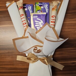 May include: A white paper bouquet with gold trim and a gold ribbon tied in a bow. The bouquet contains various chocolate bars, including Milka, Giotto, and Olio.