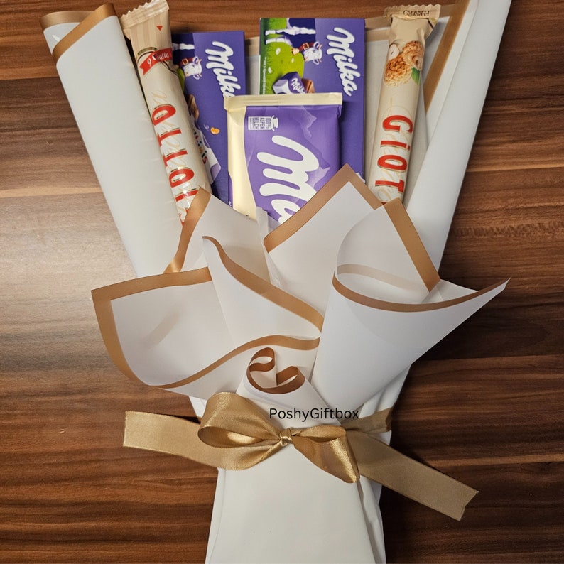 May include: A white paper bouquet with gold trim and a gold ribbon tied in a bow. The bouquet contains various chocolate bars, including Milka, Giotto, and Olio.