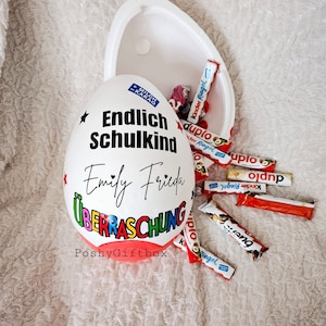Peut inclure: Un contenant blanc en forme d'&oelig;uf avec les mots "Endlich Schulkind" et "Emily Frieda" &eacute;crits dessus. Le contenant est ouvert et rempli de barres chocolat&eacute;es, dont Kinder Riegel et Duplo. Le contenant est &eacute;tiquet&eacute; "PoshyGiftbox".