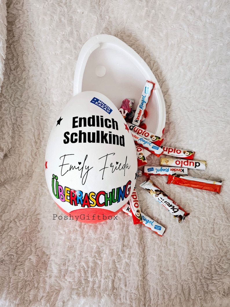 Peut inclure: Un contenant blanc en forme d'&oelig;uf avec les mots "Endlich Schulkind" et "Emily Frieda" &eacute;crits dessus. Le contenant est ouvert et rempli de barres chocolat&eacute;es, dont Kinder Riegel et Duplo. Le contenant est &eacute;tiquet&eacute; "PoshyGiftbox".