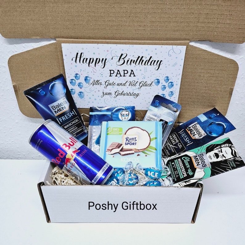 May include: A white gift box filled with various items, including a can of Red Bull energy drink, a chocolate bar, and several blue and white toiletries. The box is decorated with a blue and white card that reads "Happy Birthday Papa. Alles Gute und Viel Gluck zum Geburtstag."