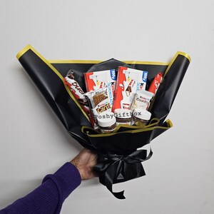 May include: A black gift bouquet with a black ribbon. The bouquet contains various chocolate bars and spreads, including Kinder, Nutella, and Cadbury.