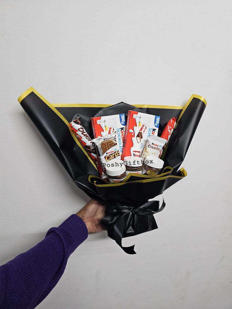 May include: A black gift bouquet with a black ribbon. The bouquet contains various chocolate bars and spreads, including Kinder, Nutella, and Cadbury.