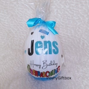 May include: A white chocolate egg with a blue ribbon and the words "Jens Happy Birthday &Uuml;berraschung PoshyGiftbox" printed on it.
