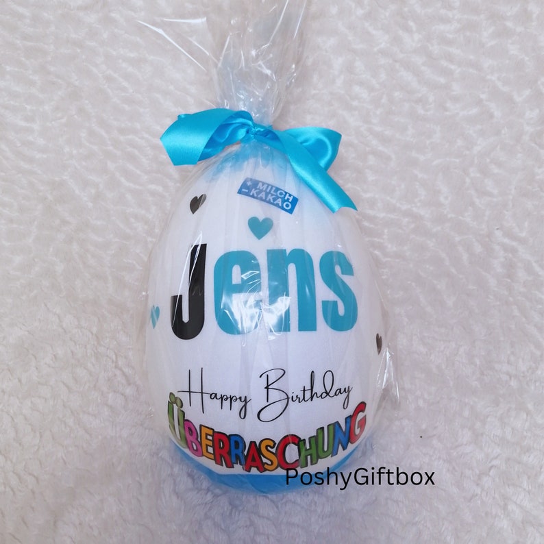 May include: A white chocolate egg with a blue ribbon and the words "Jens Happy Birthday &Uuml;berraschung PoshyGiftbox" printed on it.